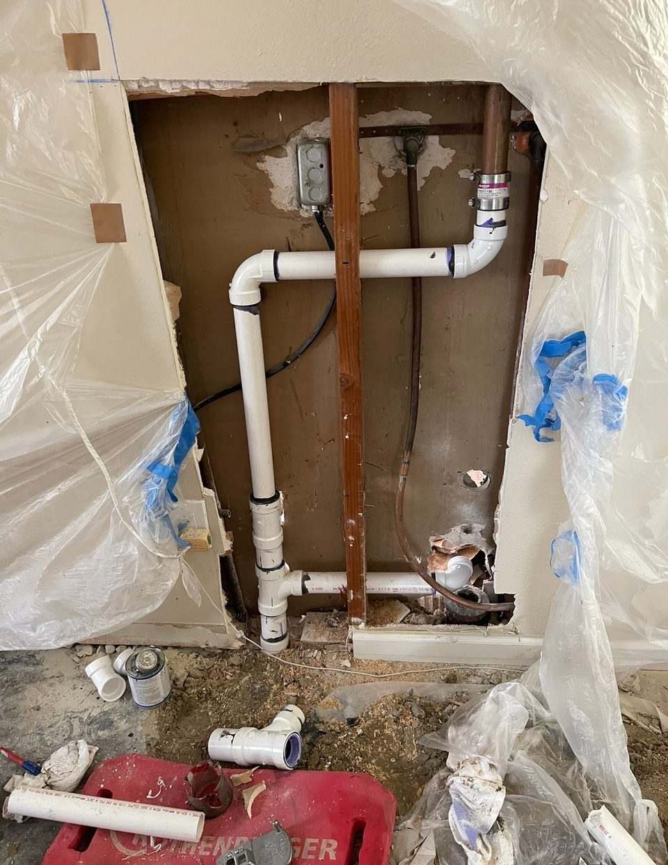 New PVC drain and vent rough-in inside an opened interior wall, with plastic sheeting hung for dust containment
