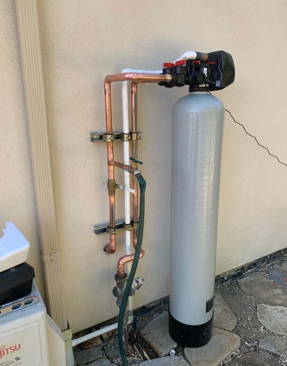 Whole-house water filtration tank connected with copper piping and a bypass valve against a stucco exterior wall