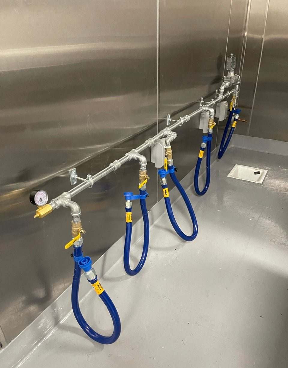 Blue commercial gas connectors and shutoff valves running along a stainless steel commercial kitchen wall