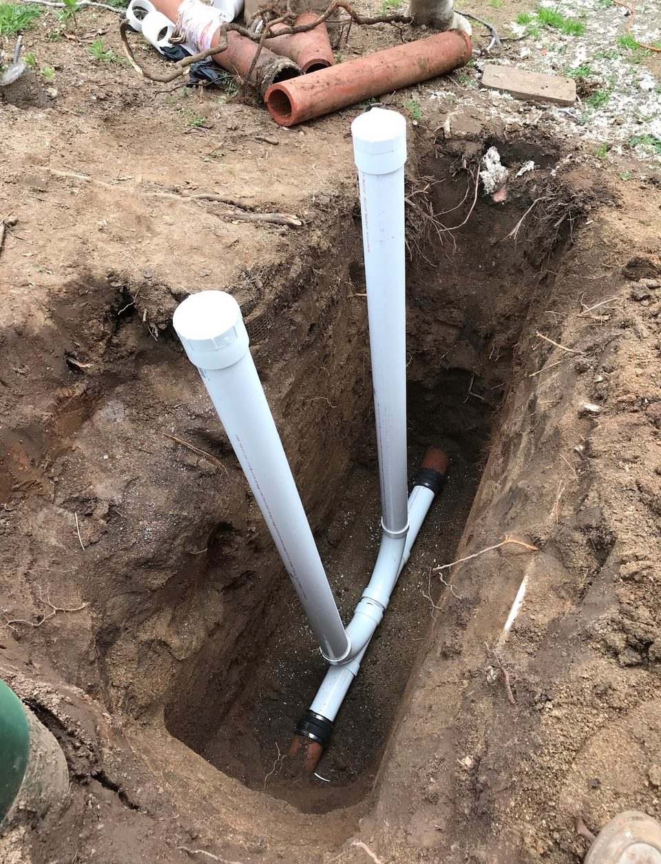 Two white PVC cleanout risers set into an open trench, with sections of removed cast iron pipe stacked at the edge