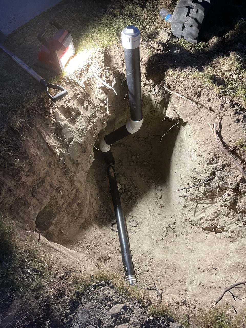 Black ABS sewer line with a white cleanout riser installed in an open trench, photographed at night under work lights