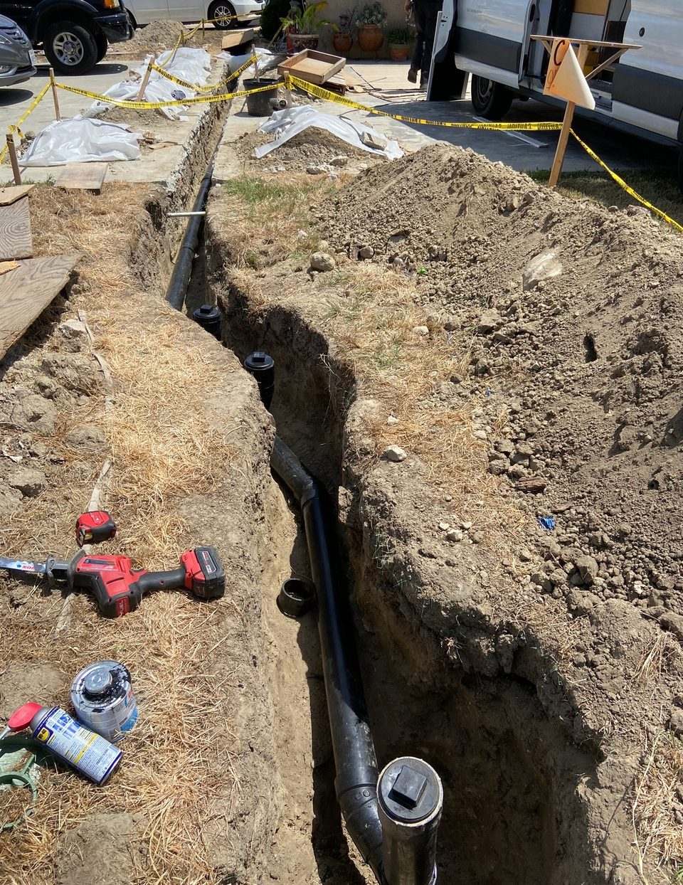 Long open trench running along a driveway with a new black ABS sewer line and cleanout fitting, cordoned off with caution tape