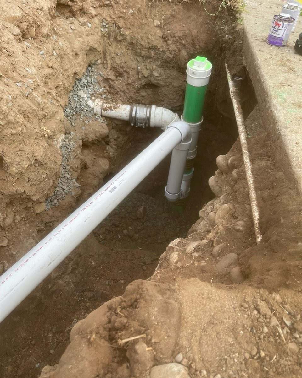 New PVC sewer line and green-capped cleanout installed in a trench alongside a building foundation