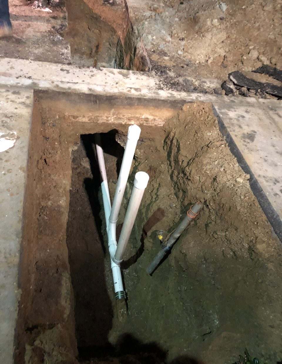 New white PVC drain lines rising from a trench cut through a concrete slab, photographed at night