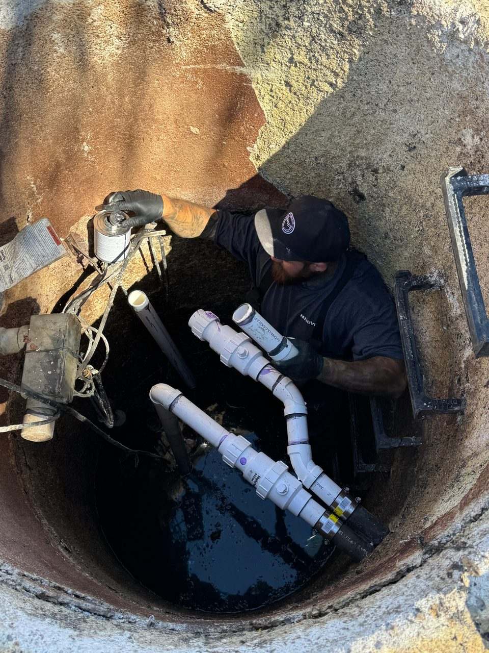Plumber working inside a below-grade concrete vault, installing new PVC piping above standing water