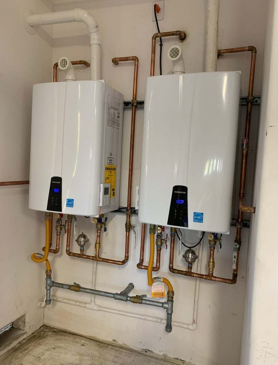 Two white Navien tankless water heaters mounted side by side on a wall, connected with copper manifolds, isolation valves and yellow gas lines